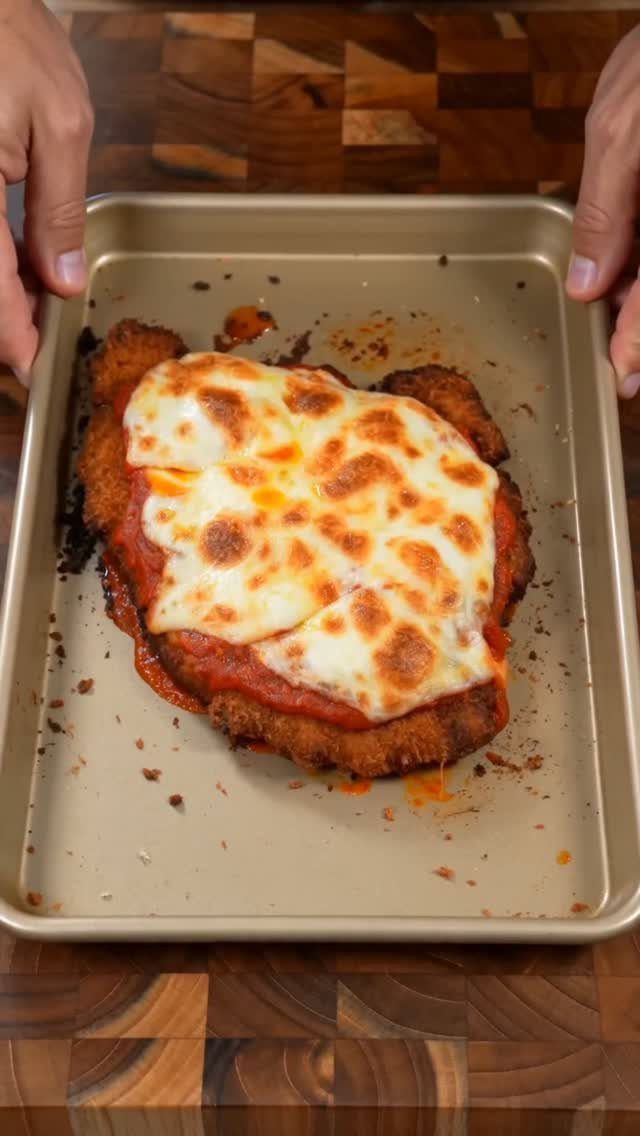 Breaded chicken parmigiana with melted cheese on a baking sheet over wooden board.