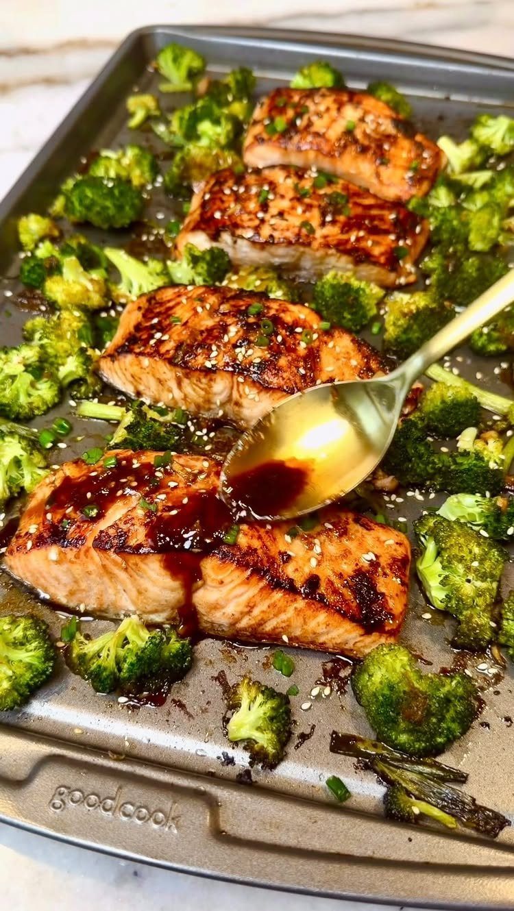 Glazed salmon fillets and roasted broccoli on a baking sheet with sauce being poured.