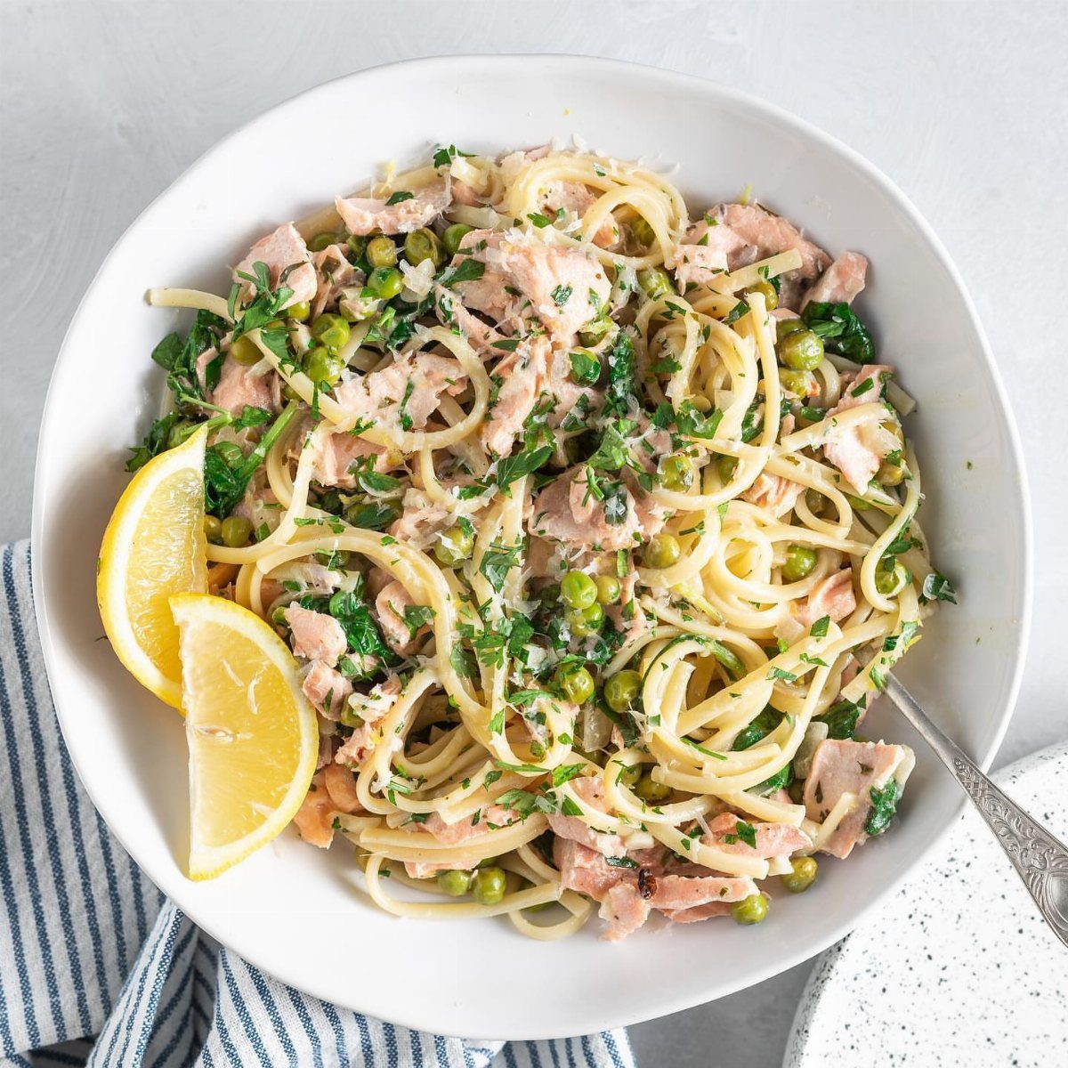 Linguine pasta with flaked salmon and green peas served with lemon wedges.
