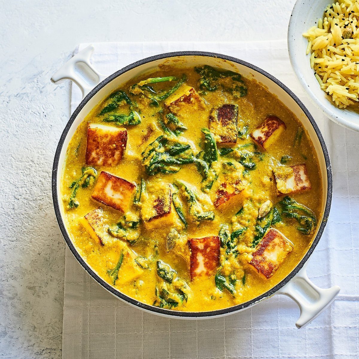 Yellow curry with paneer and spinach in a white pot with side rice.