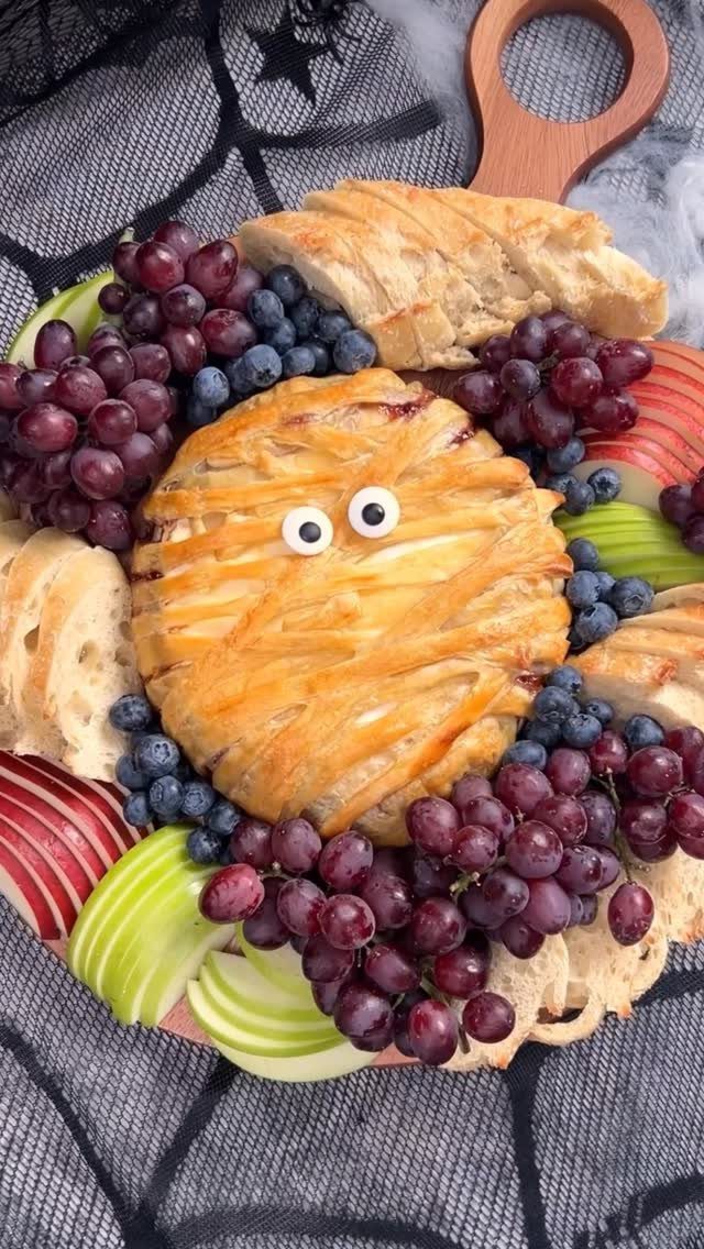 Baked brie with edible eyes surrounded by grapes, blueberries, apples, and baguette slices.
