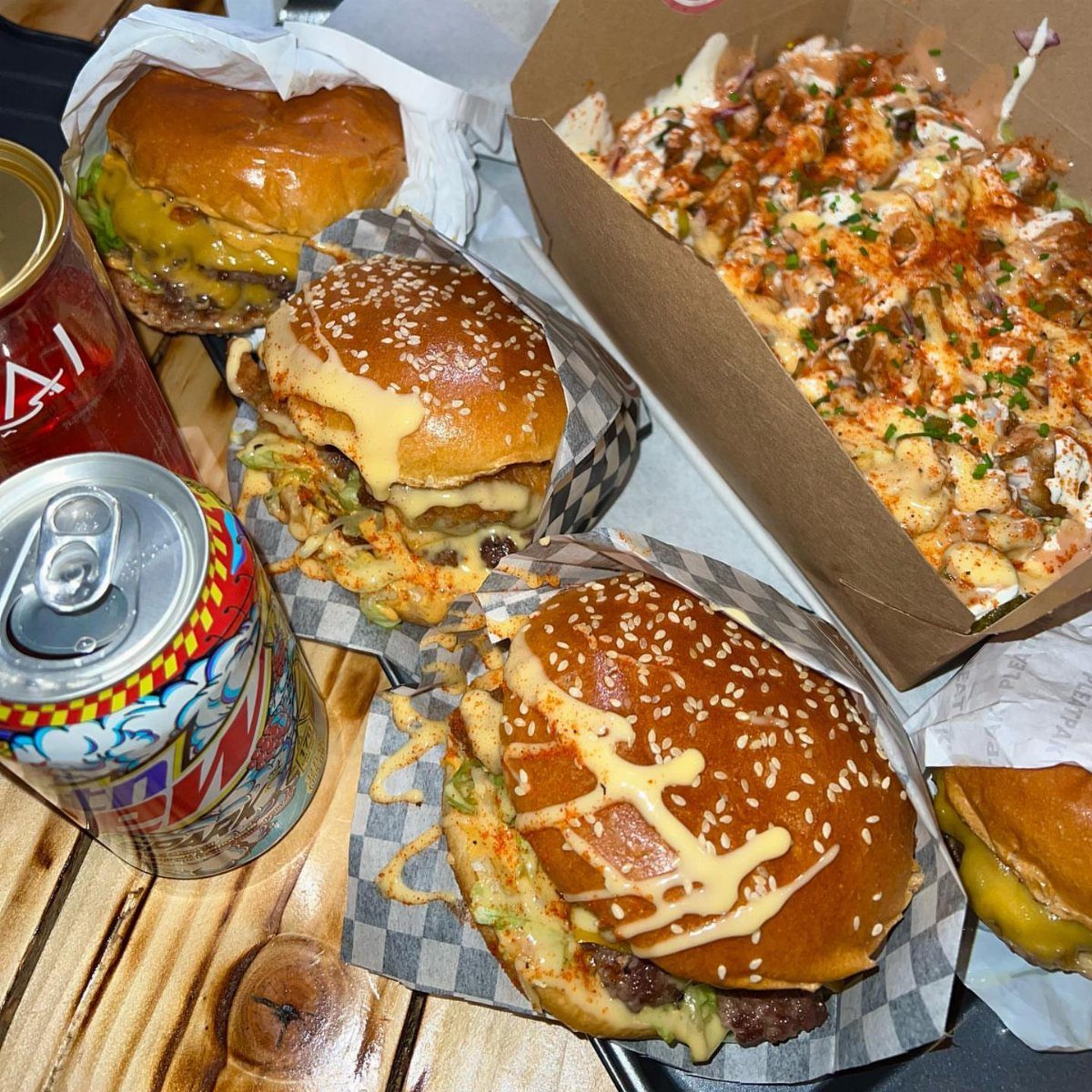 Cheeseburgers and loaded fries with beverage cans on a rustic wooden table.