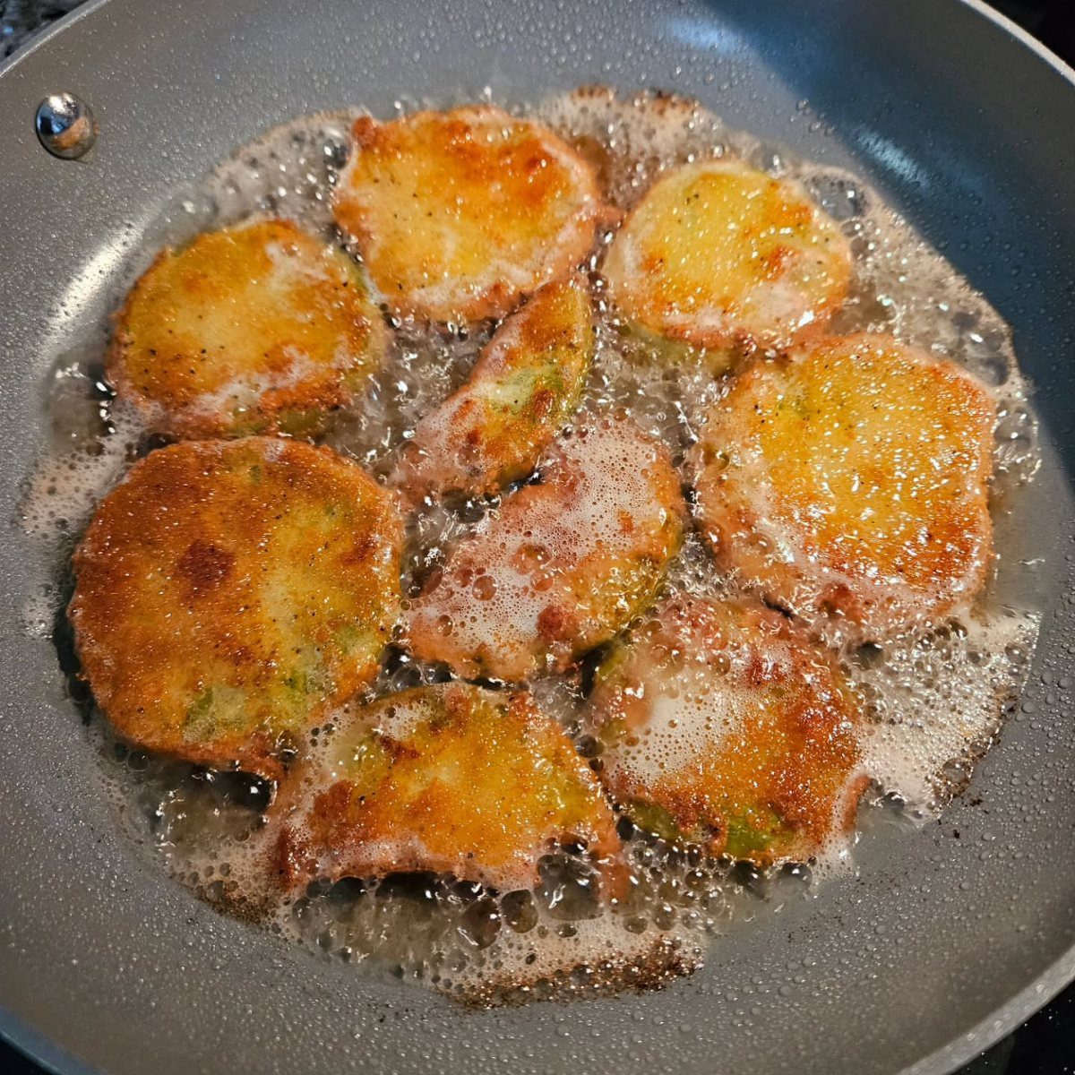 Breaded green tomato slices frying in a grey skillet with bubbling hot oil.