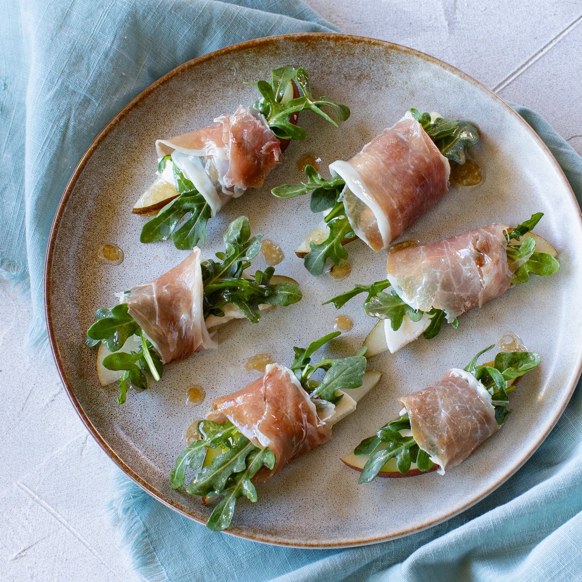 Prosciutto-wrapped pear slices with arugula and honey on a ceramic plate.