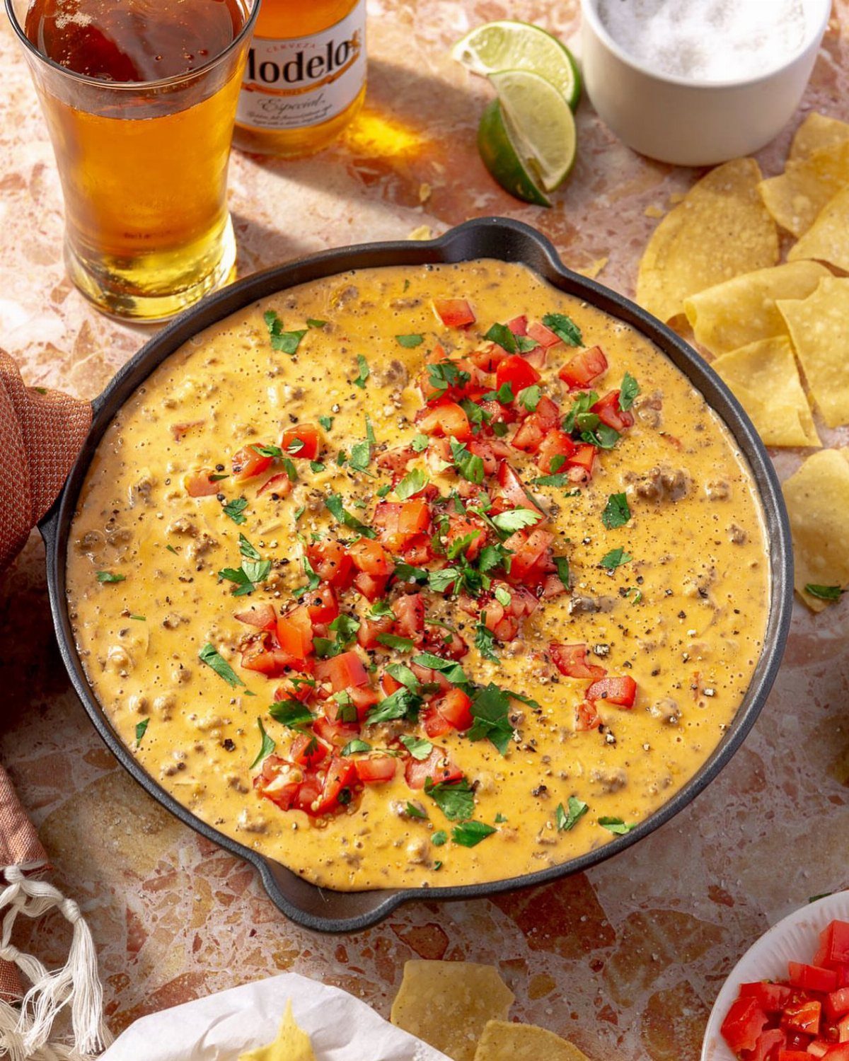 Queso dip in a cast-iron skillet with tortilla chips and beer on marble.