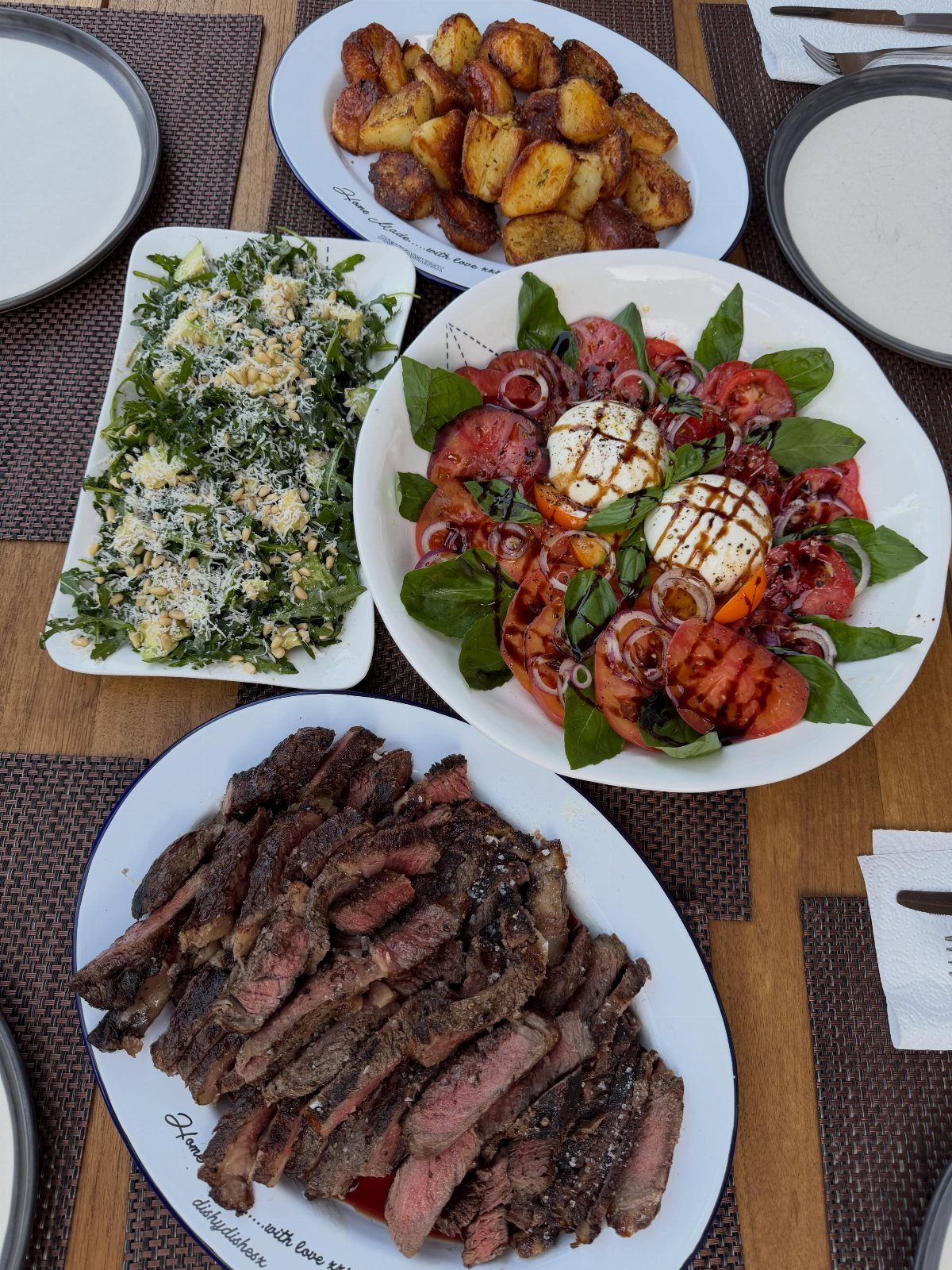 Grilled steak, Caprese salad, green salad, and roasted potatoes on a wooden table.