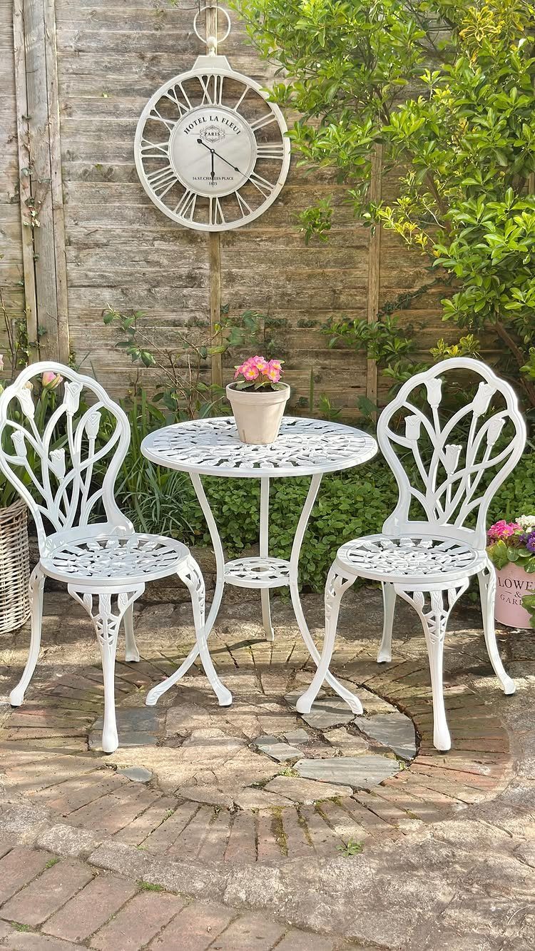 White ornate metal bistro table and chairs on a brick patio with garden foliage