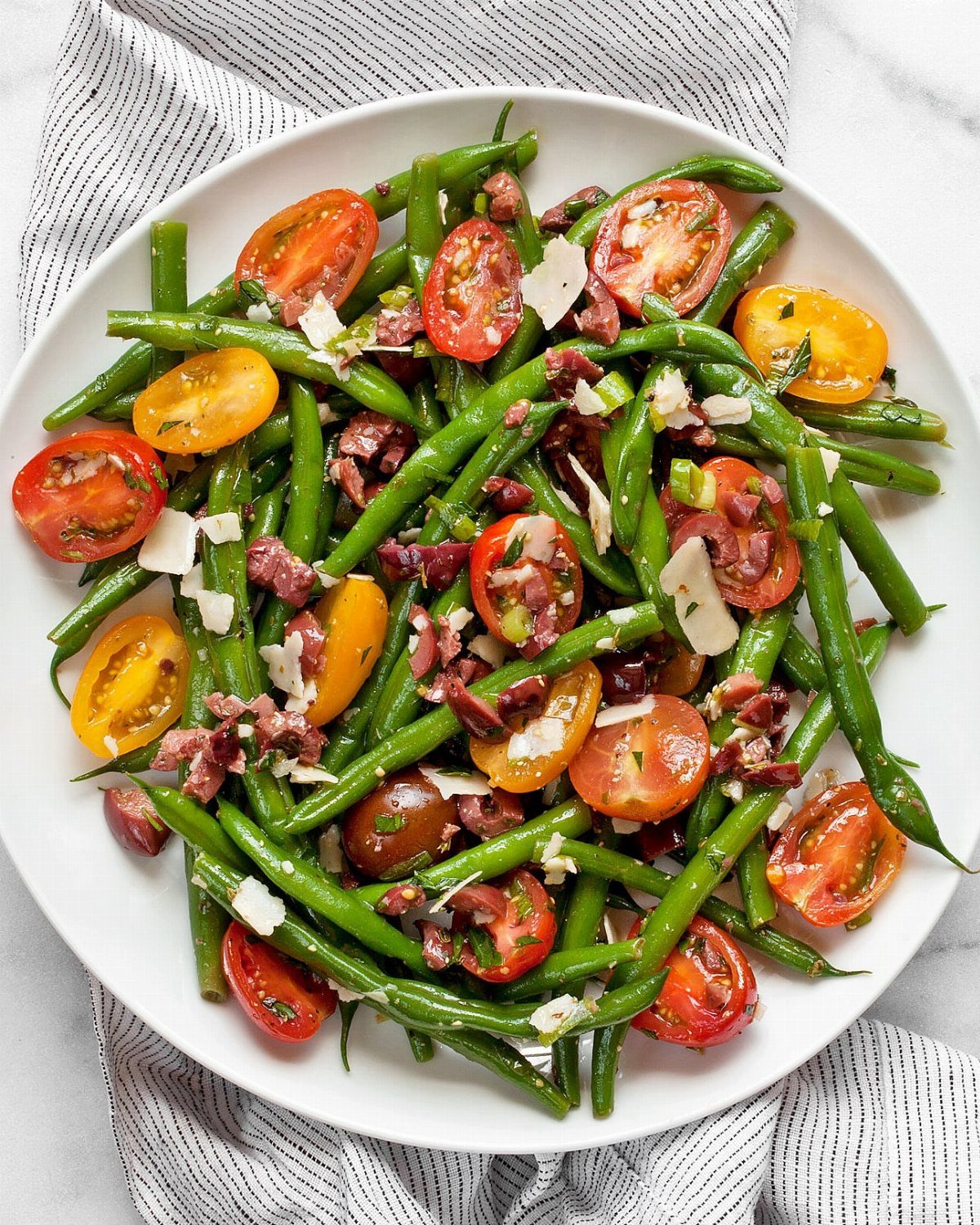Green bean salad with cherry tomatoes, cured meat, and parmesan on a white plate.