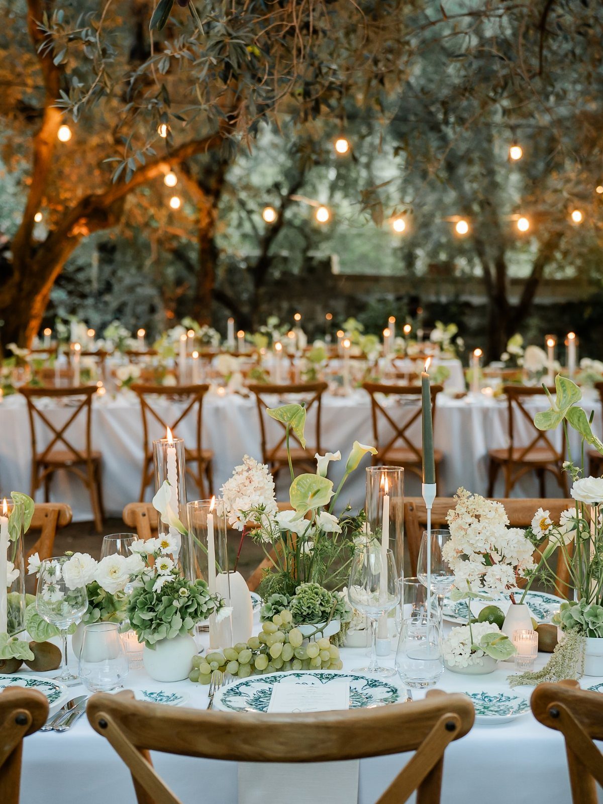 Elegant outdoor dinner table set with white linens, flowers, and hanging string lights.
