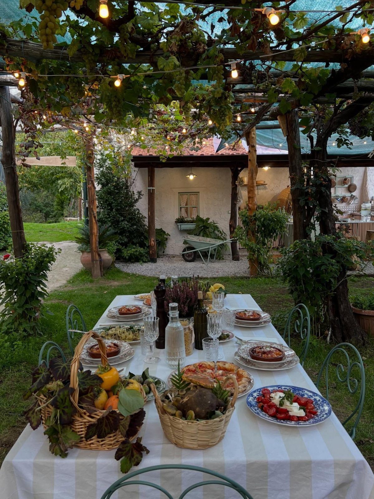 Outdoor dinner table with Italian food set under a lit grape trellis garden.