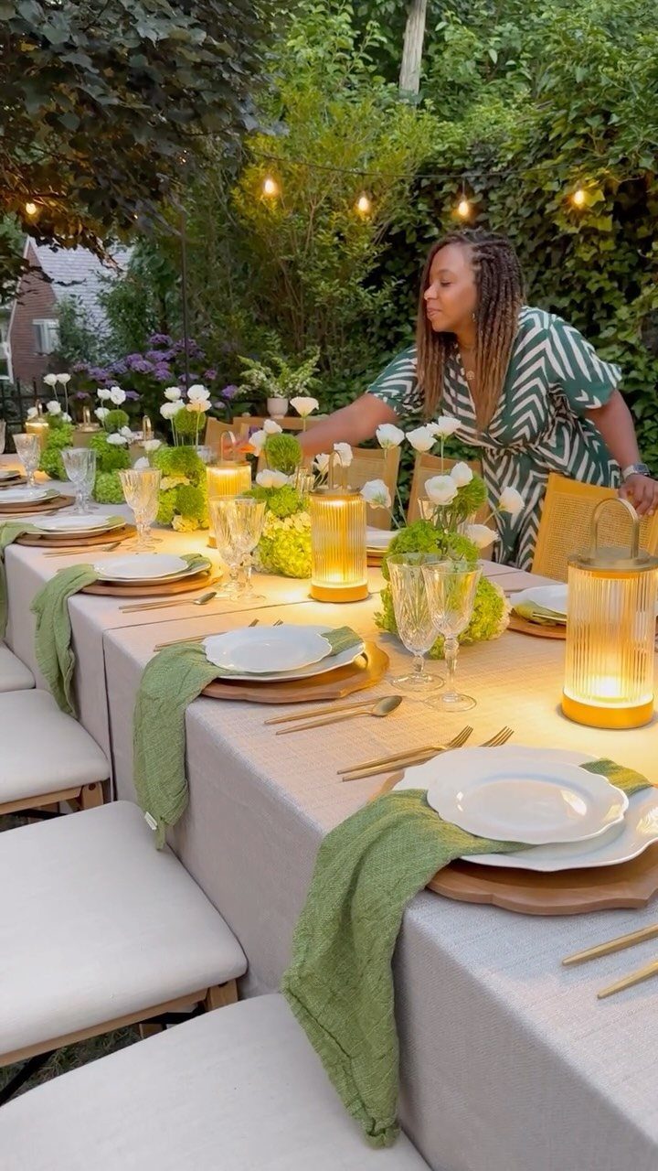 Woman arranging a decorated outdoor dining table with green linens and glowing lanterns.