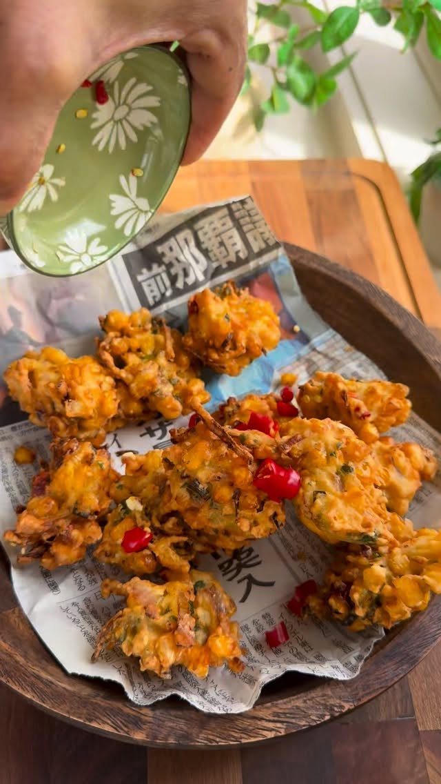 Golden-brown corn fritters with red chilies in a wooden bowl on newspaper paper.
