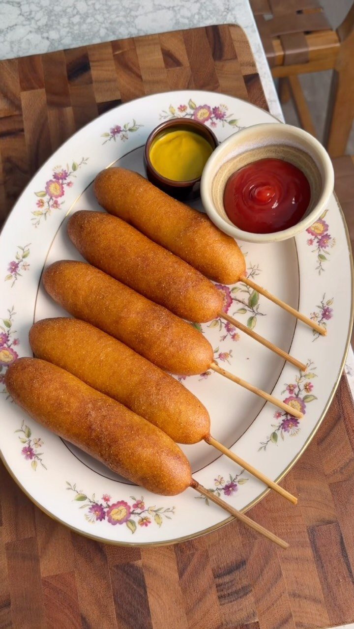 Corn dogs on a floral platter with mustard and ketchup on wooden board