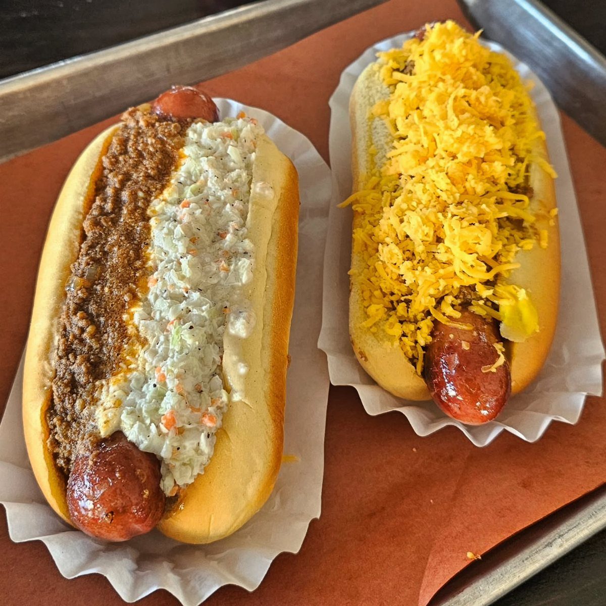 Two loaded hot dogs with chili, coleslaw, and shredded cheese on a tray.