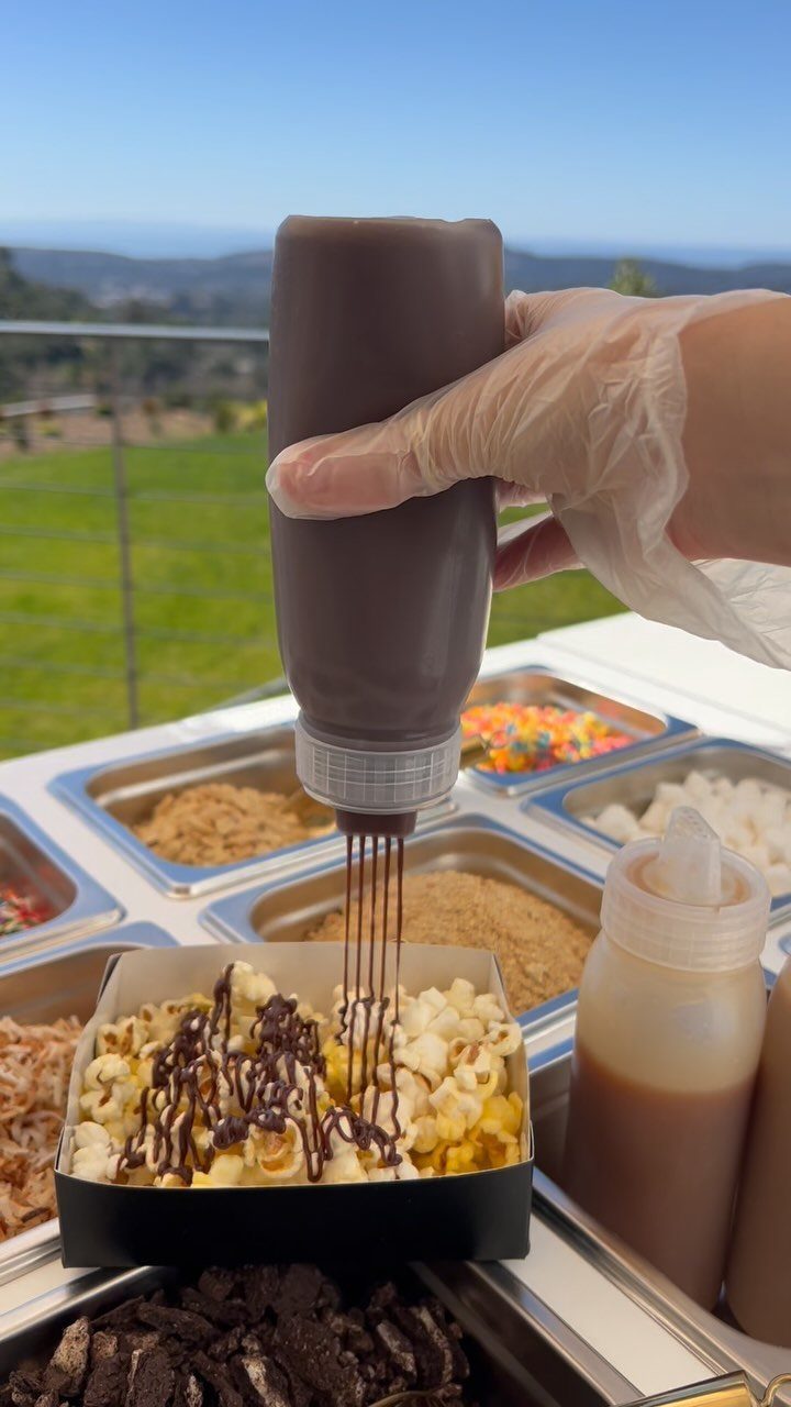 Popcorn in a black container being drizzled with chocolate sauce at a station.