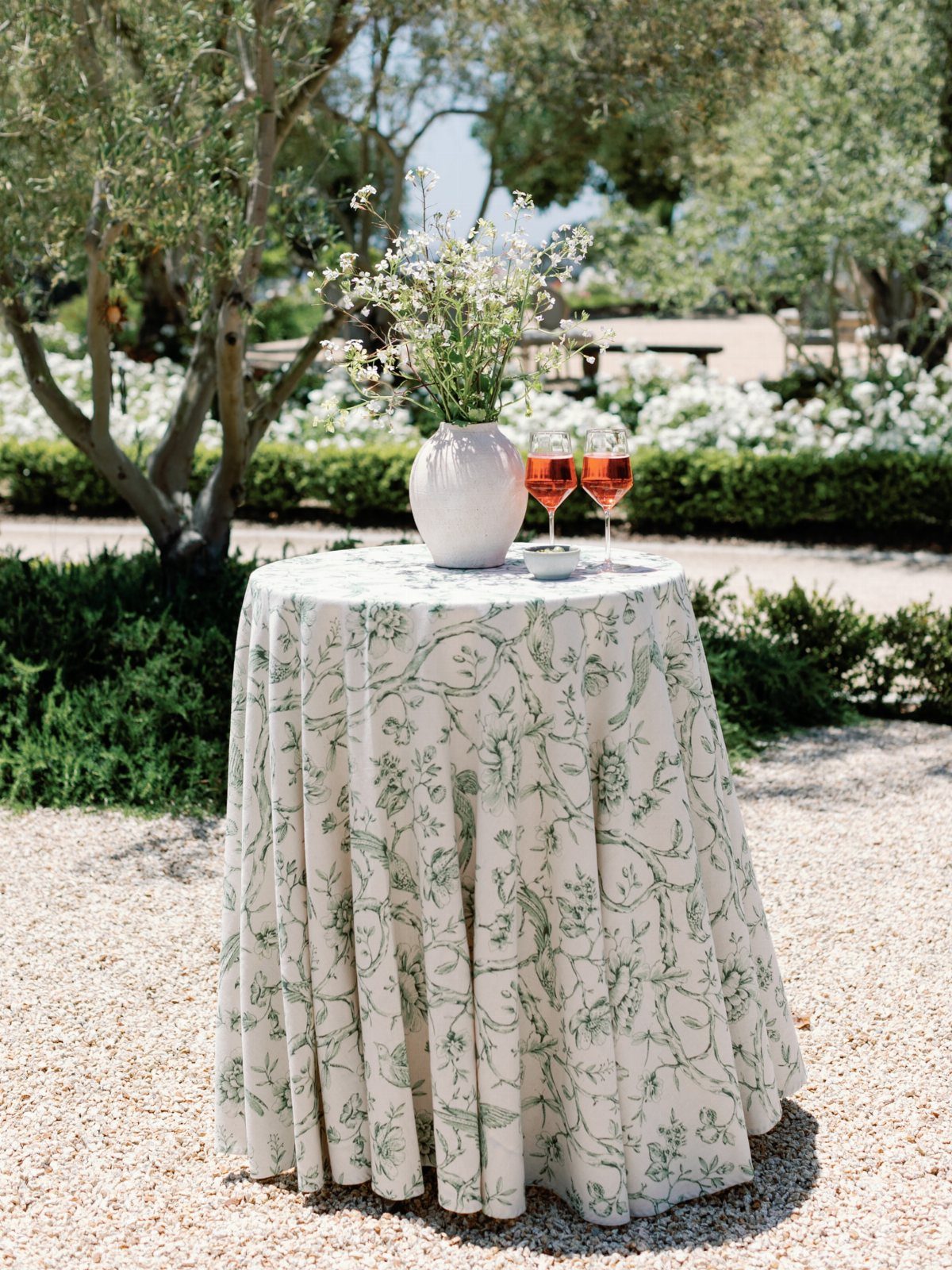 Round table with botanical print tablecloth, white flower vase, and two wine glasses.