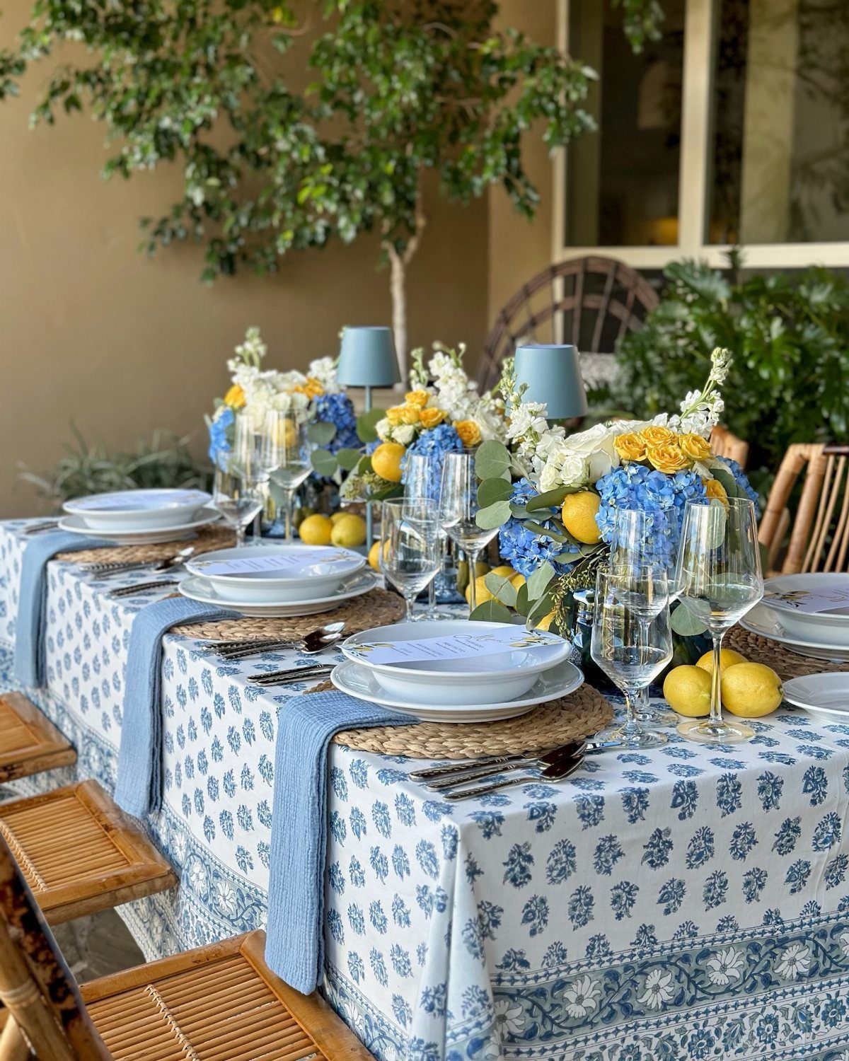 Table setting with blue patterned cloth, woven placemats, white plates, and floral centerpieces.