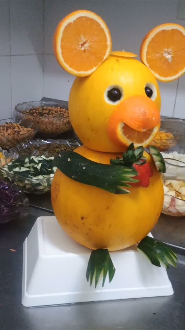 Melon mouse sculpture with orange slice ears and cucumber limbs on white base