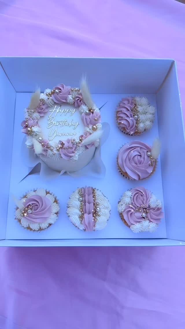 Small cake and cupcakes with purple and white frosting in a white box.