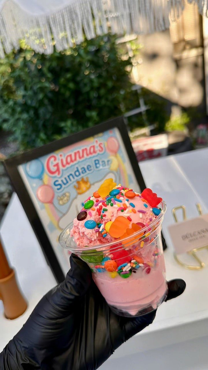 Pink ice cream sundae with colorful toppings held in front of a sundae bar sign.