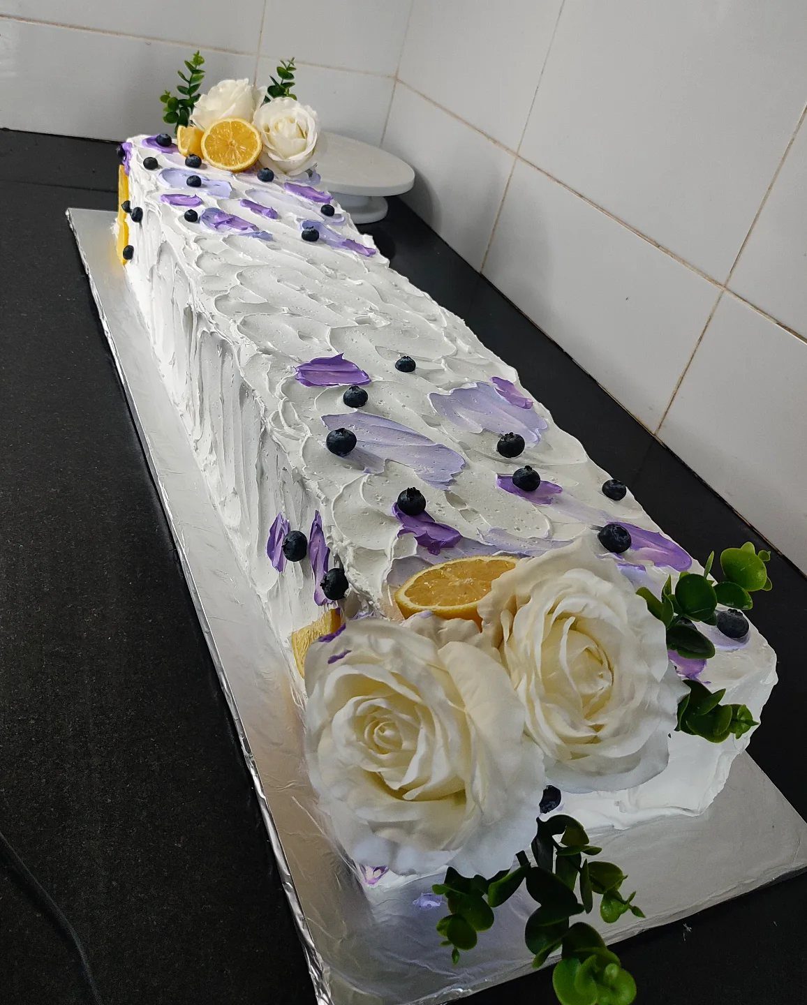 Rectangular white frosted sheet cake decorated with blueberries, lemon slices, and white roses.