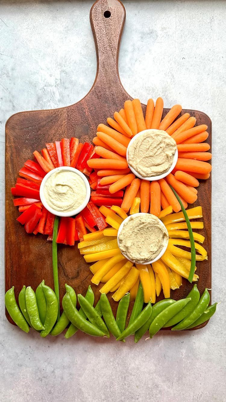 Vegetable platter with bell pepper flowers and hummus dip on a wooden board.