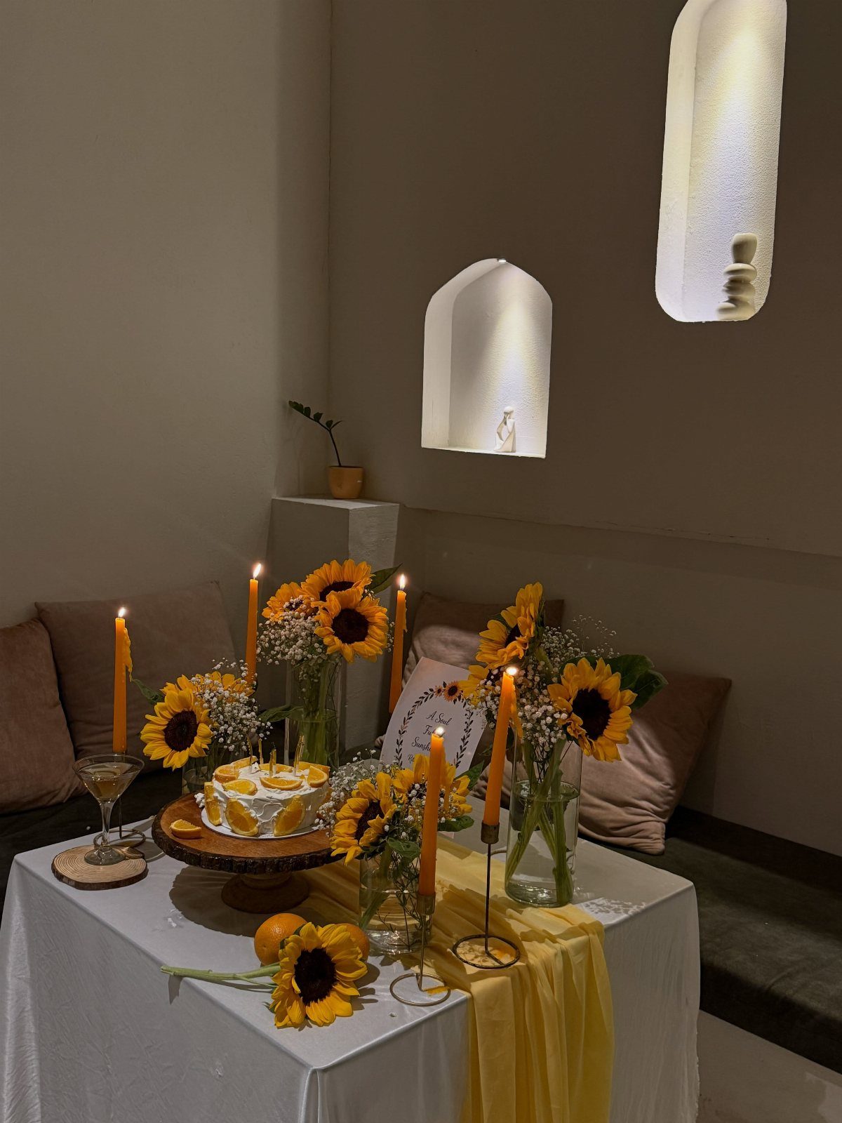 Cake on a wooden stand surrounded by yellow sunflowers and orange taper candles.