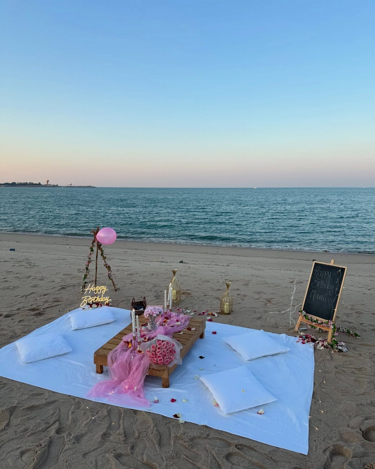 Beach birthday picnic setup with a low wooden table, pink decor, and white pillows.