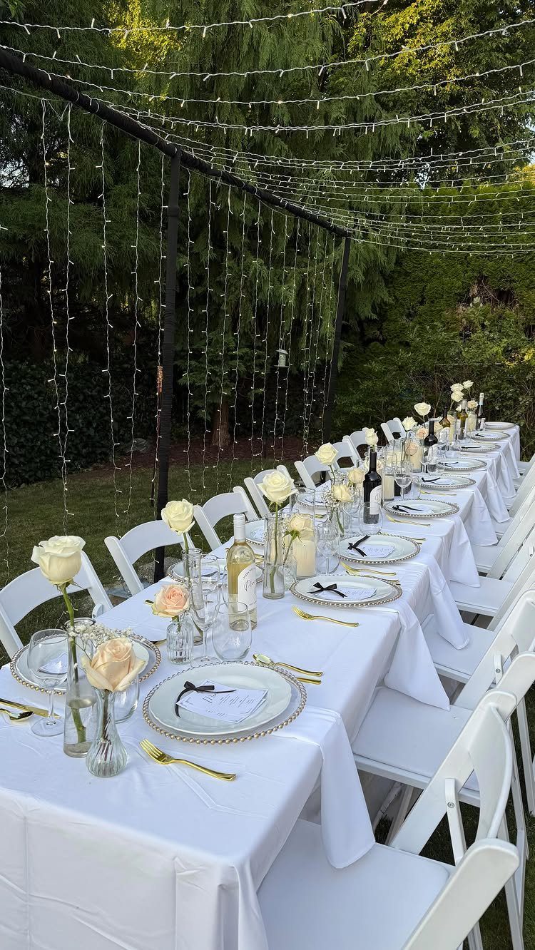Long outdoor dining table with white linens, white roses, and overhead string lights.