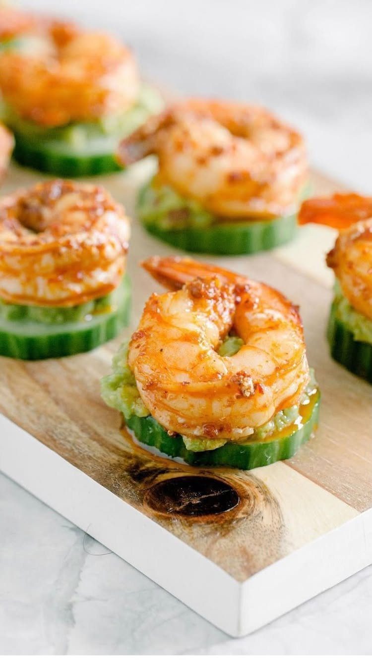 Cooked shrimp with guacamole on cucumber slices arranged on a wooden board.