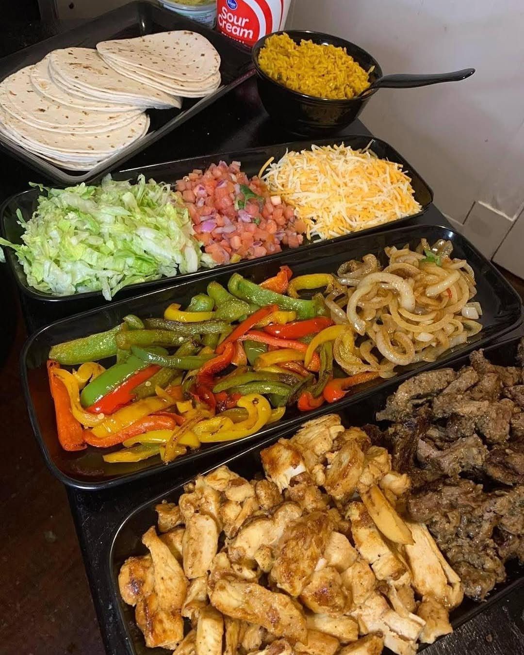 Taco bar spread with grilled meats, vegetables, tortillas, and various toppings in trays.