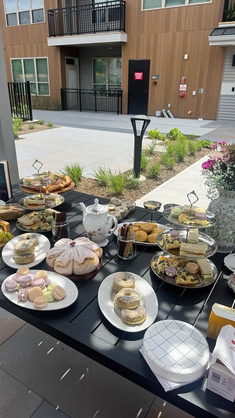 Assorted tea sandwiches and pastries on tiered stands with a white teapot outdoors.