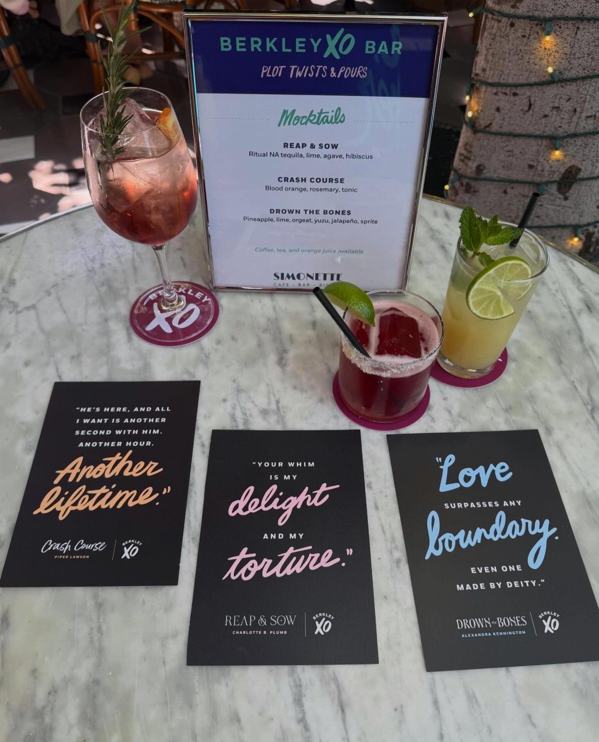 Three colorful cocktails and mocktails on a marble table with a themed menu.