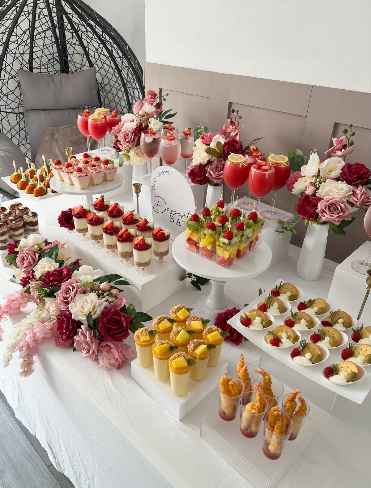 Dessert table with fruit cups, mini cakes, red cocktails, and red and white roses.