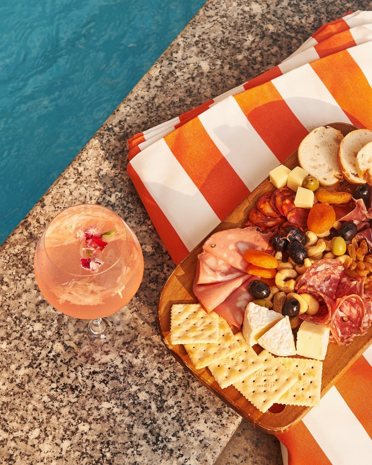 Pink spritz cocktail and charcuterie board on granite poolside with orange striped towel