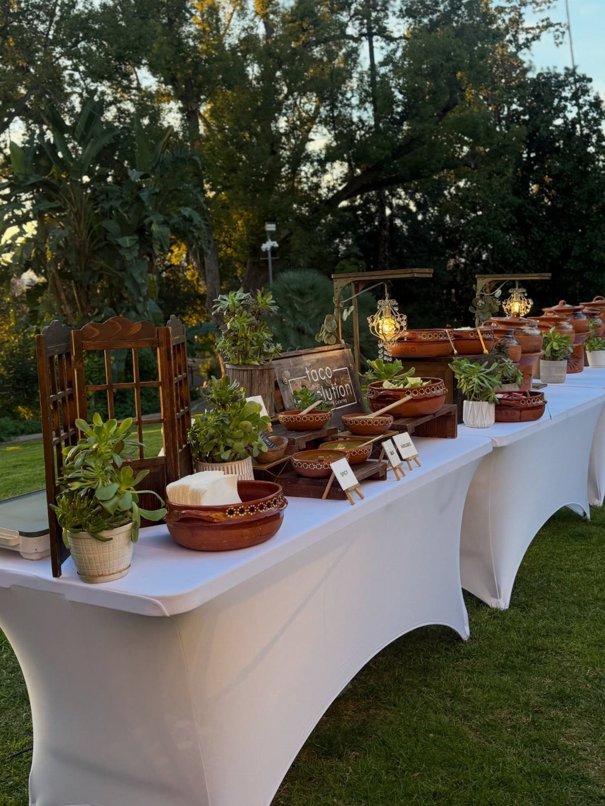Taco station with bowls of ingredients and tortillas on white tablecloths outdoors.