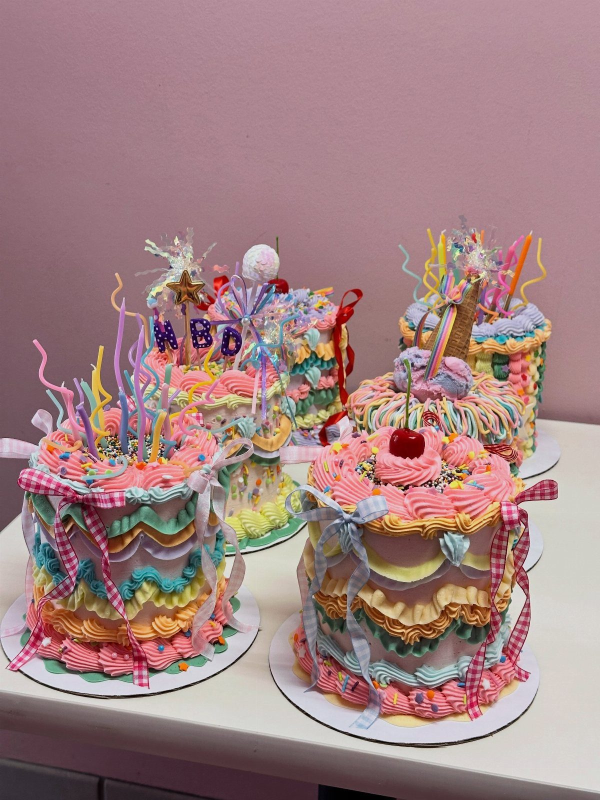 Five colorful maximalist cakes with ribbons, sprinkles, and frosting on a white table.