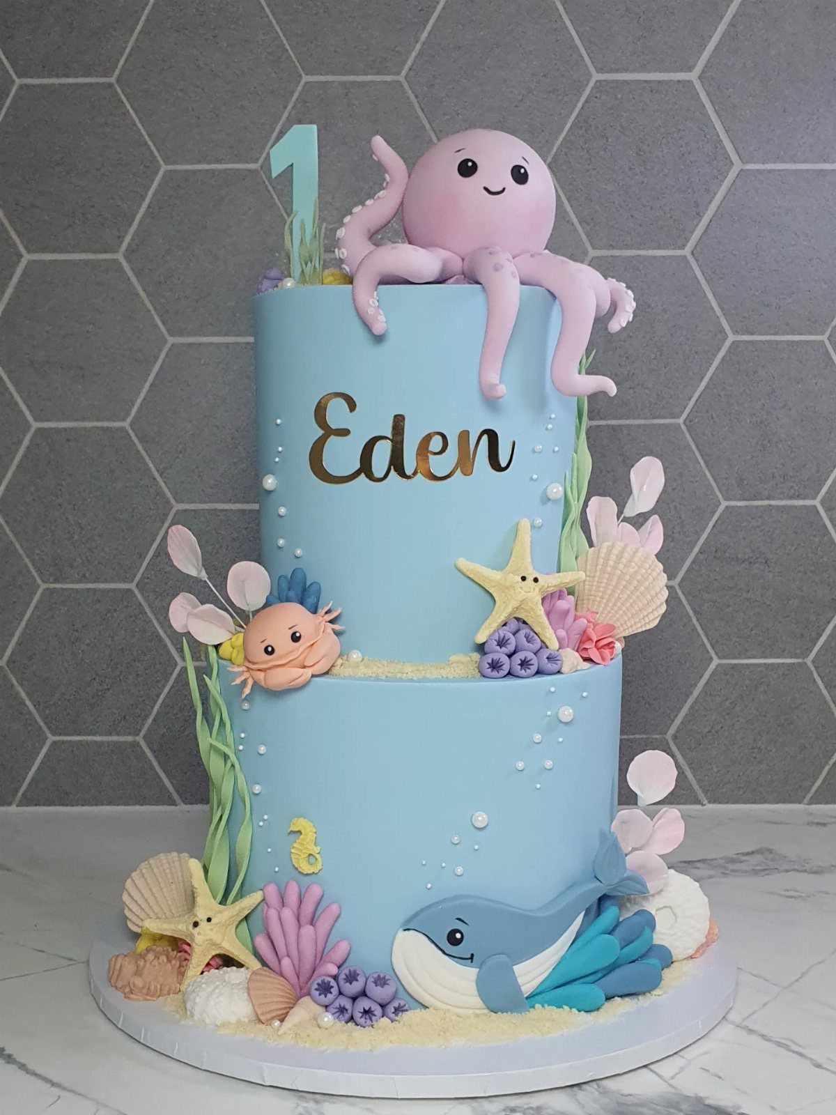 Two-tier blue ocean themed cake with pink octopus, blue whale, and seashells.