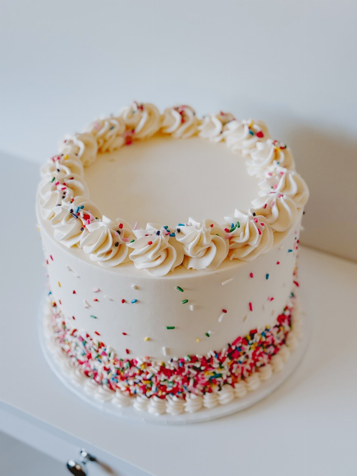 White frosted cake with colorful sprinkles around the base and piped cream swirls.