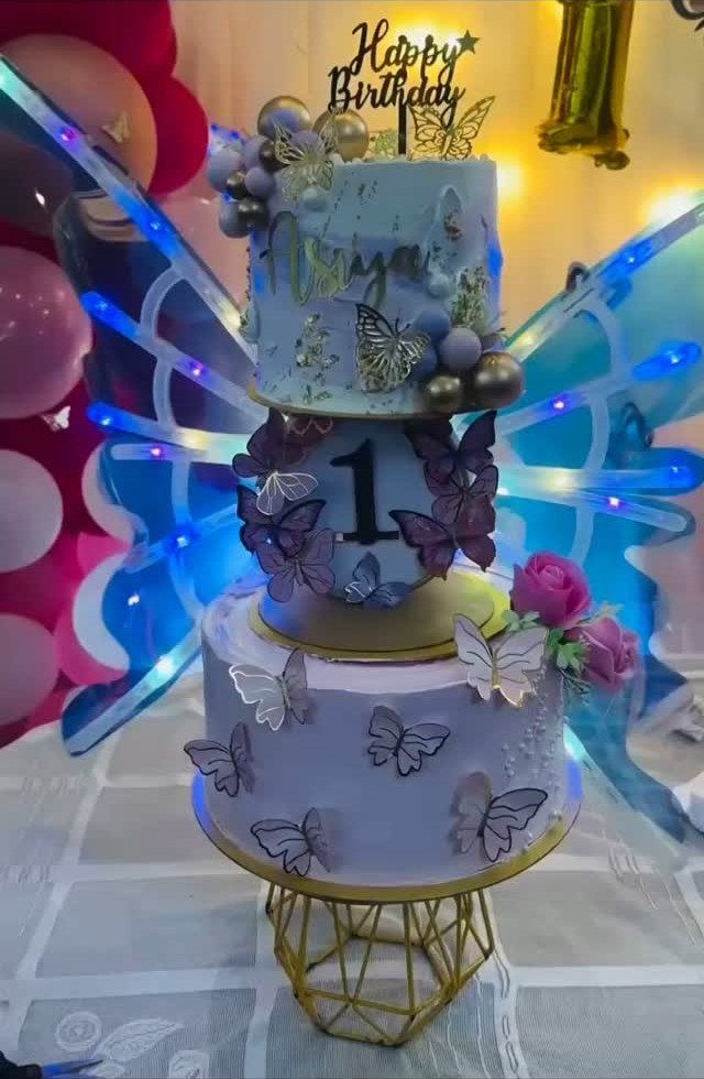Three-tiered blue birthday cake with butterflies on a gold geometric wire cake stand.