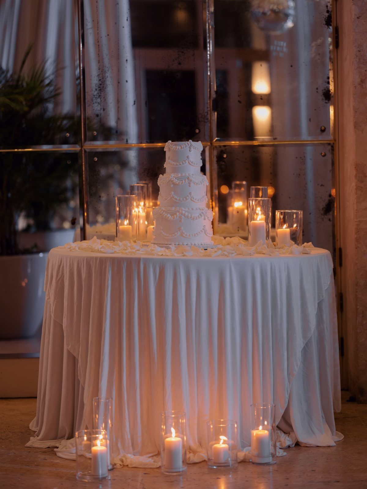 White tiered wedding cake on a draped table surrounded by glowing cylinder candles.