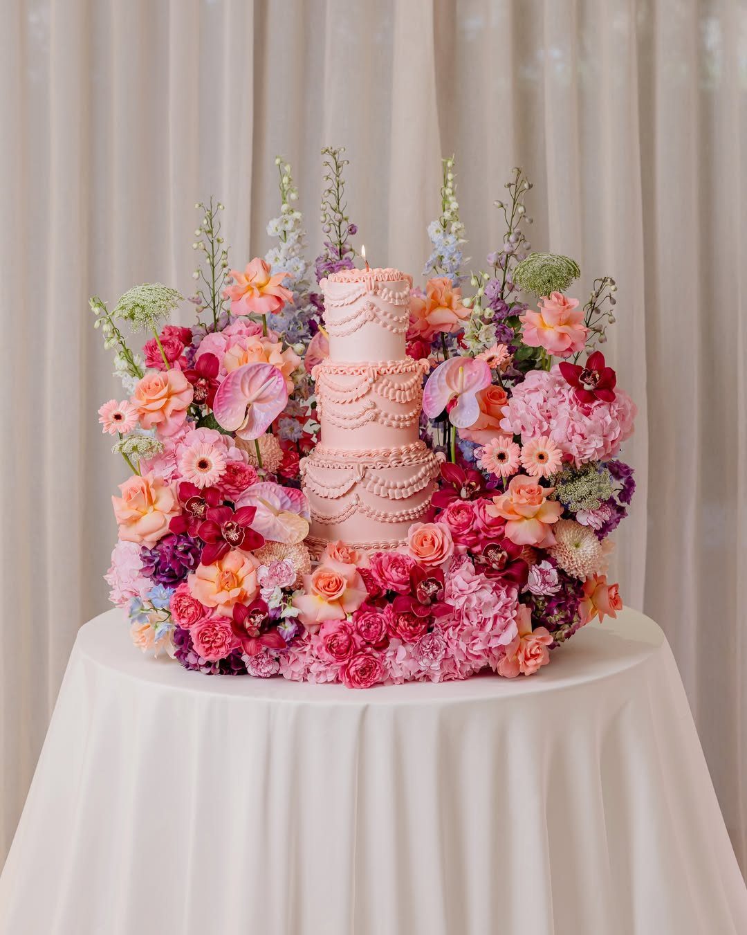 Pink tiered cake surrounded by a lush meadow of colorful flowers on white table