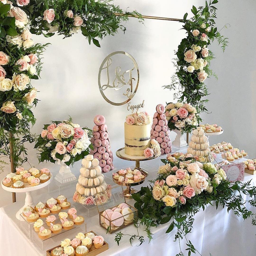 Dessert table with tiered cake, donuts, and pink roses under a floral arch.