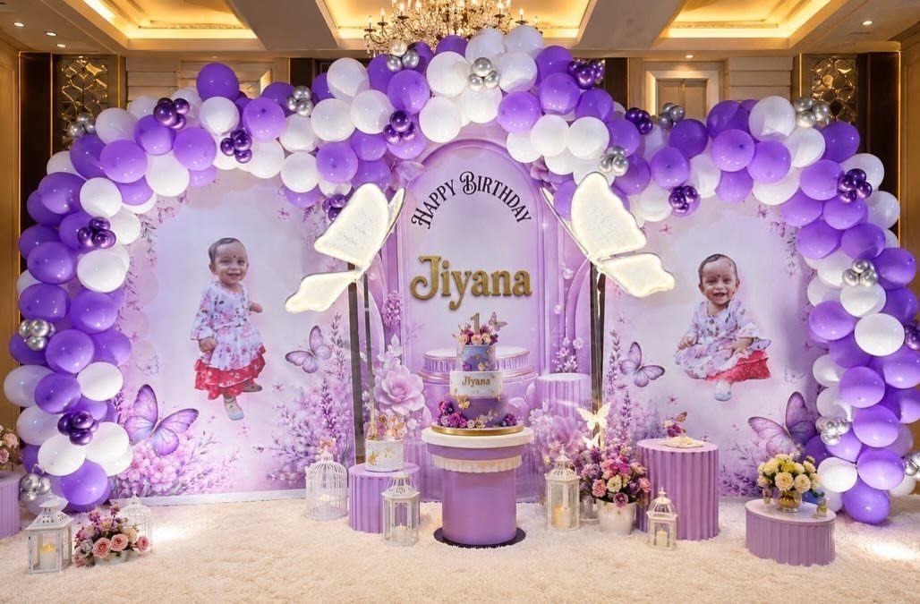 Purple and white balloon arch framing a birthday cake table with baby photos.
