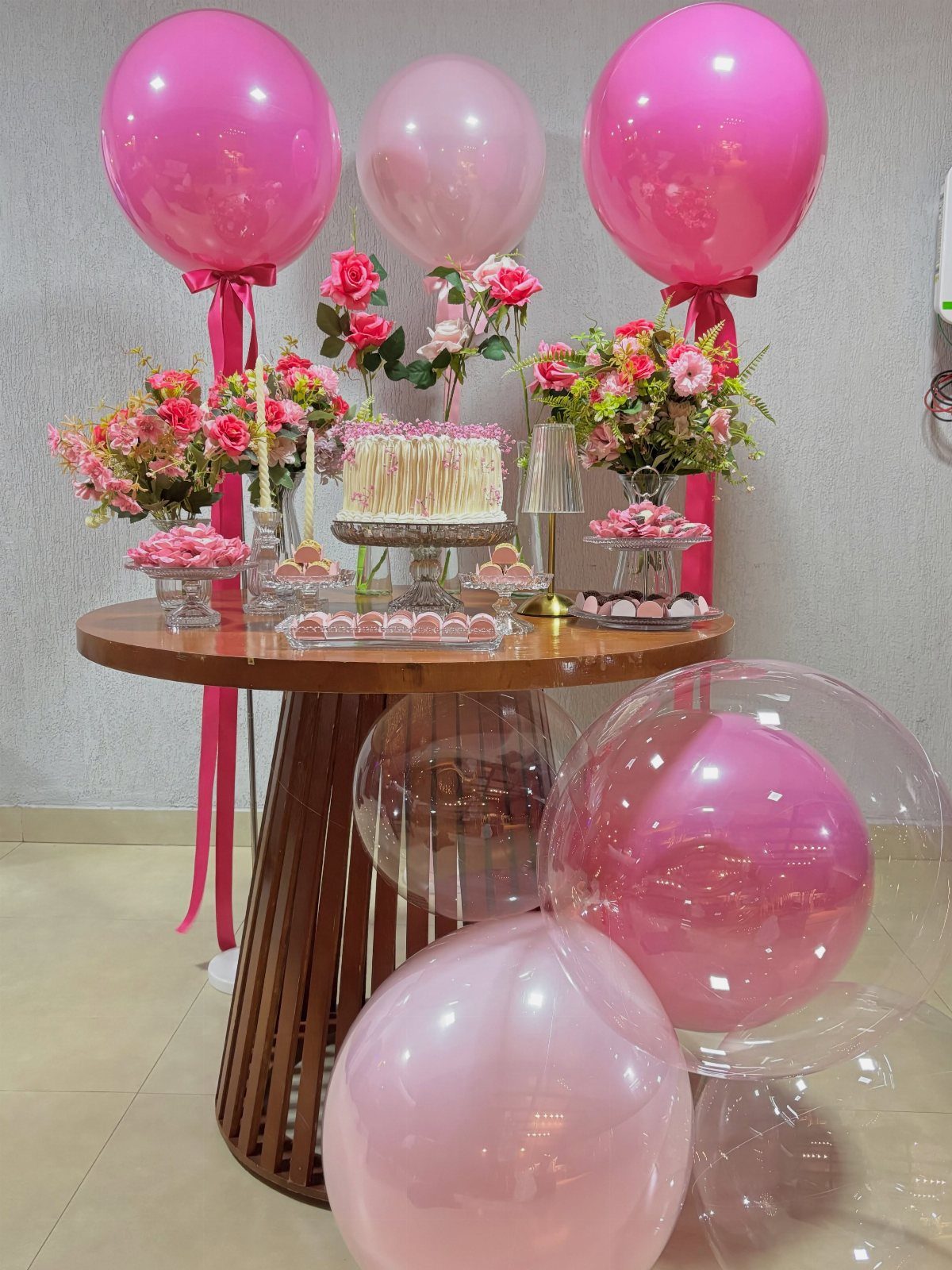 Round wooden cake table decorated with pink balloons, floral arrangements, and a white cake.