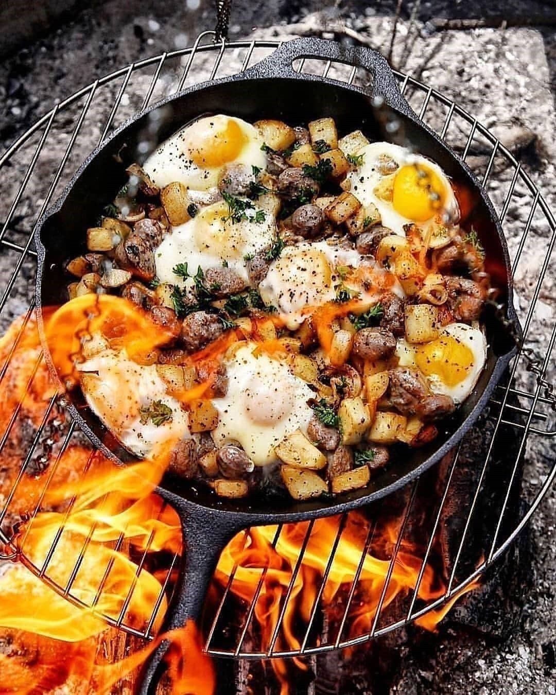 Cast iron skillet with eggs and potatoes cooking over an open campfire flame.