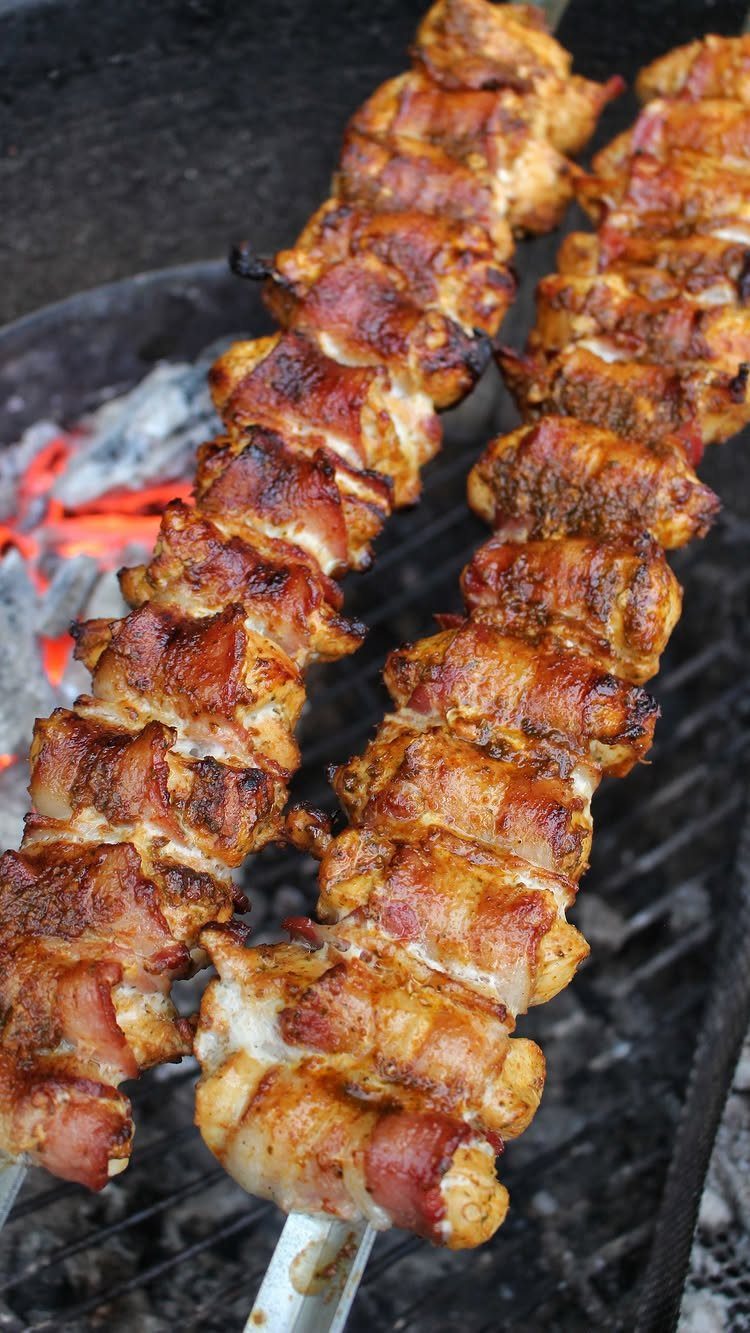 Bacon-wrapped meat skewers grilling over glowing orange campfire embers on a metal grate