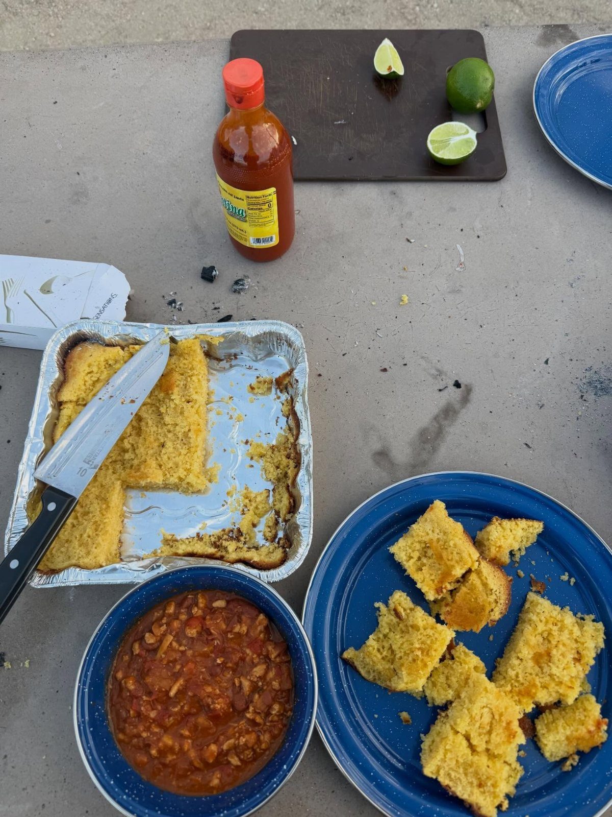 Bowl of hearty chili with cornbread, hot sauce, and lime on outdoor table.