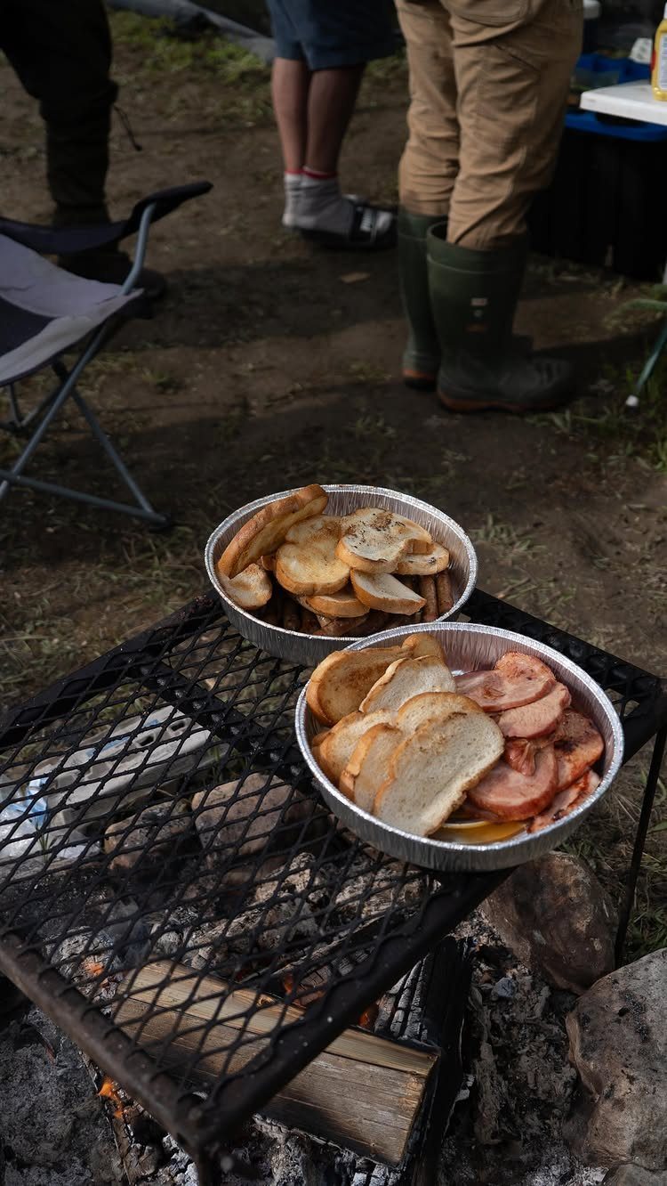 Sliced sausages and toasted baguette in foil trays on a campfire grill.