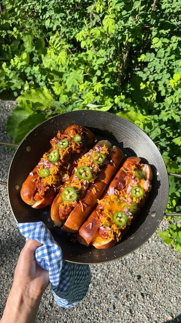 Chili cheese dogs with jalapeños in a black pan held outdoors.